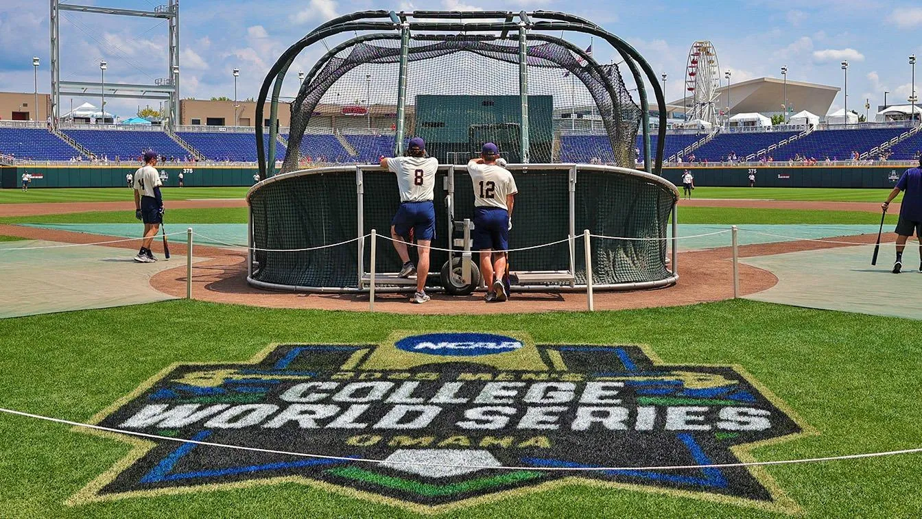 Baseball players in white and navy uniforms behind batting cage with College World Series logo on grass and Ferris wheel in background