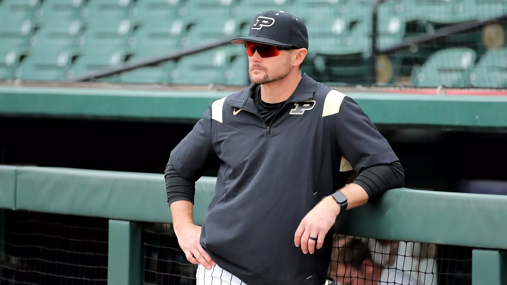 Baseball coach in black Purdue cap and jacket with gold accents leaning on dugout fence wearing sunglasses